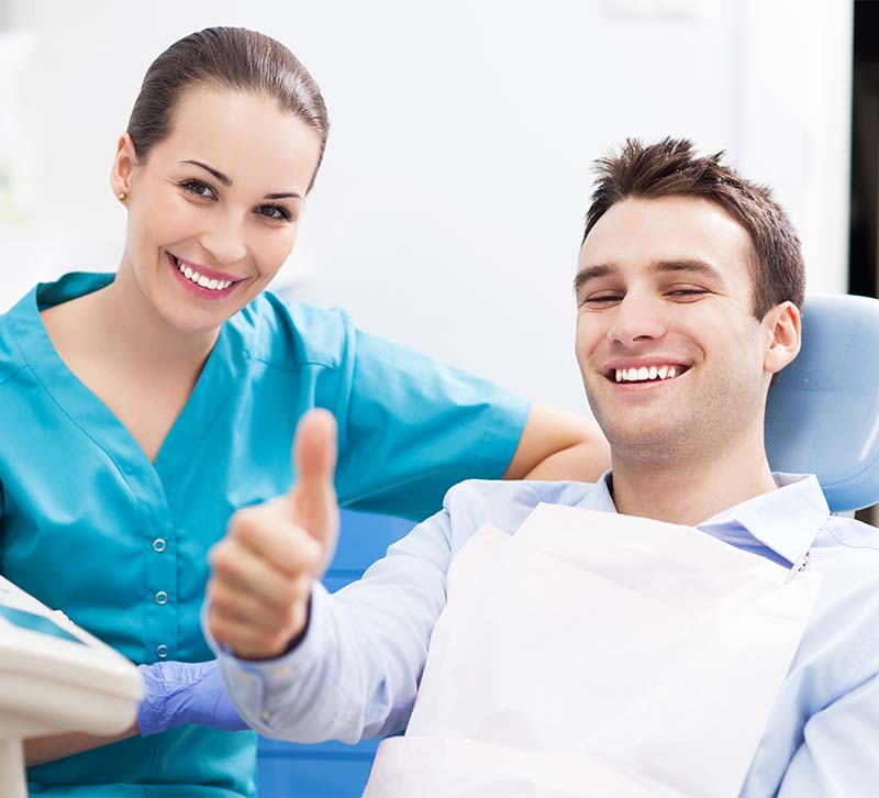 A dental professional and a patient in a dental office, both smiling and giving thumbs up.