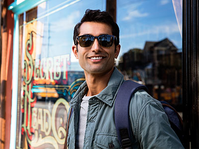A young man in sunglasses, standing outside a storefront, wearing a backpack and a jacket.